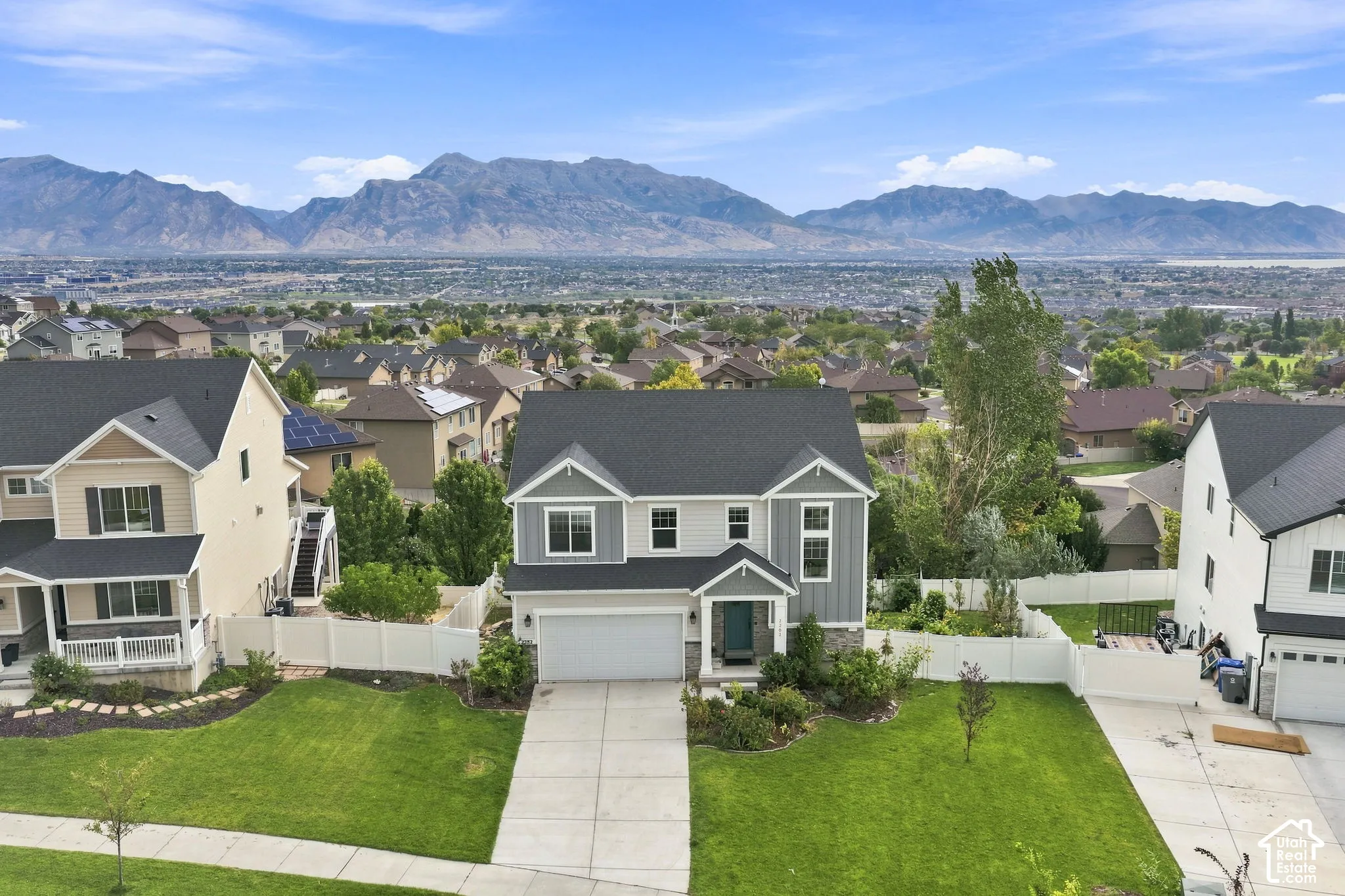 Two-story residence featuring a mixed-material exterior of horizontal lap siding and vertical board-and-batten, a front-facing attached garage, a concrete driveway, a white vinyl privacy fence, and an expansive mountain backdrop
