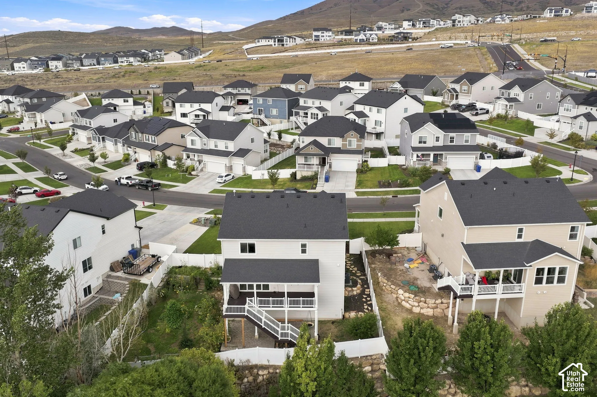 Aerial perspective of a multi-level suburban residence featuring a rear elevated deck with white railing, a stone-accented retaining wall, a white vinyl privacy fence, and mountain scenery