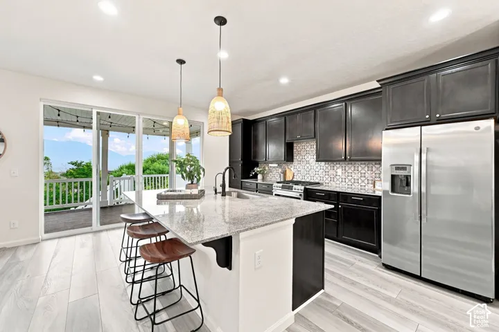 Kitchen featuring dark cabinetry, stone-surface island with integrated sink, patterned tile backsplash, stainless steel appliances, and wood-finish flooring