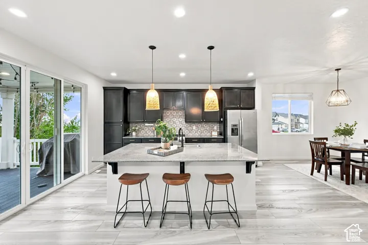 Kitchen featuring a large central island with light-toned stone countertop, dark custom cabinetry, patterned tile backsplash, wood-finish flooring, and pendant lighting