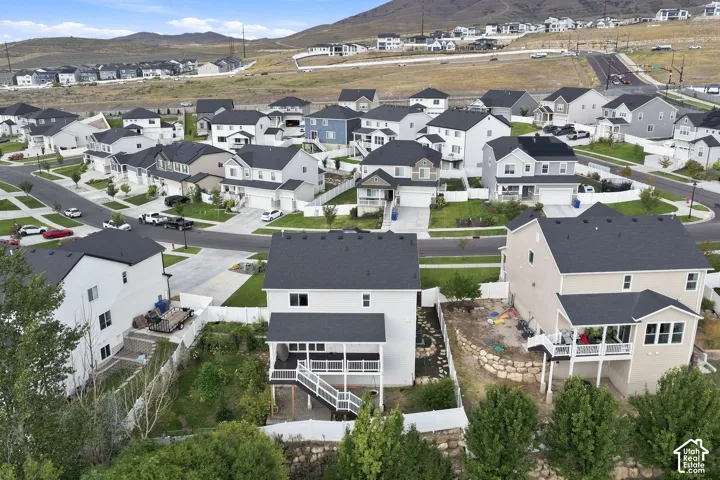 Aerial perspective of a multi-level suburban residence featuring a raised wooden deck with a staircase, stone retaining wall, white vinyl fencing, and mountain terrain backdrop