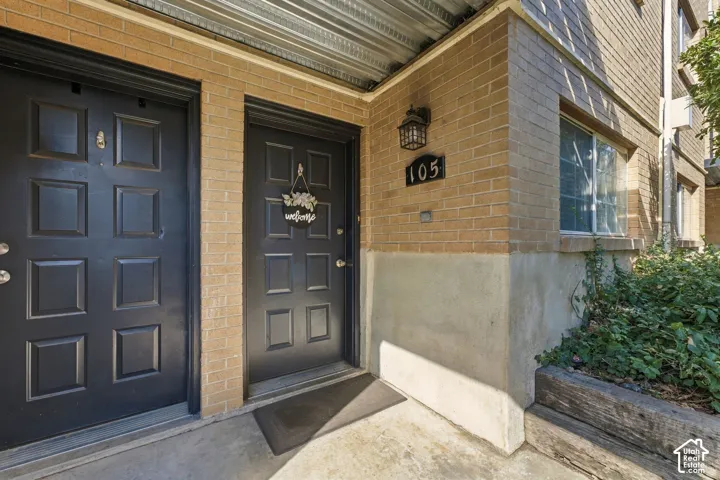 Exterior entryway featuring dark-toned paneled doors, tan brick cladding, and a wall-mounted lantern-style light fixture