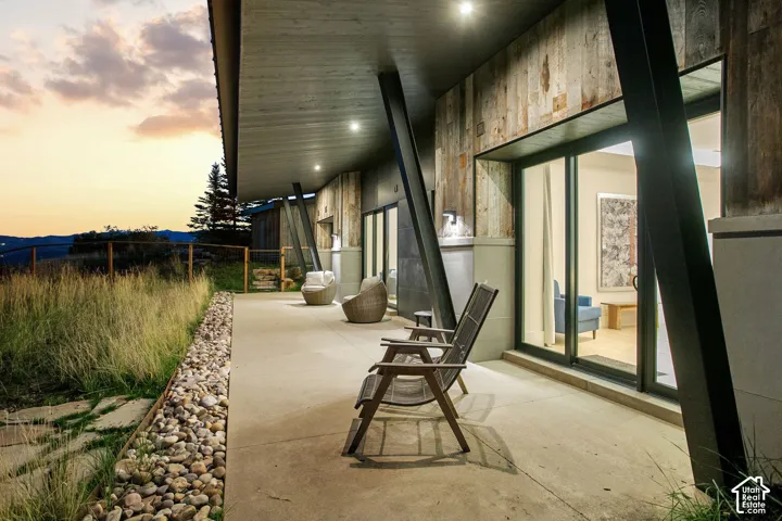 Covered concrete patio featuring angular structural support beams, wood-plank siding, large sliding glass doors, and river rock landscaping bordering the natural terrain