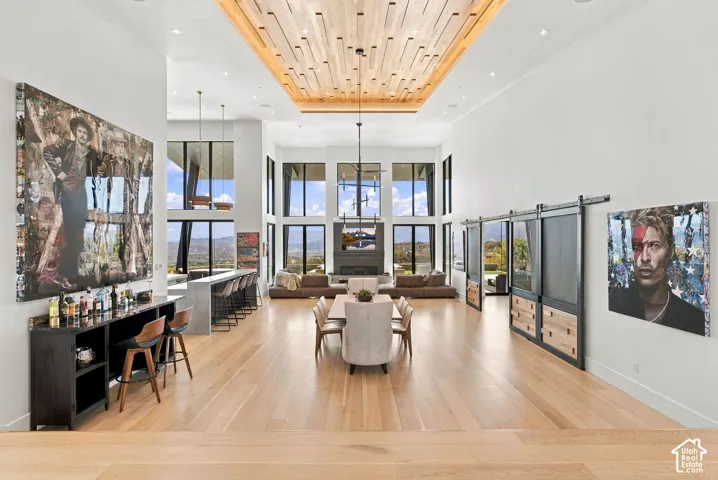 Expansive open-concept living area featuring custom wood-slat tray ceiling, floor-to-ceiling windows, light wood-finish flooring, bar, and sliding metal-framed barn doors