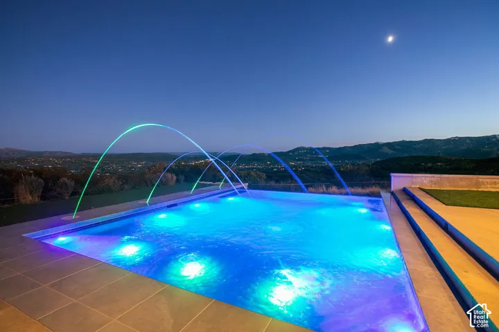 Infinity-edge swimming pool featuring integrated water jets, submerged lighting, neutral-toned tile decking, and mountain backdrop