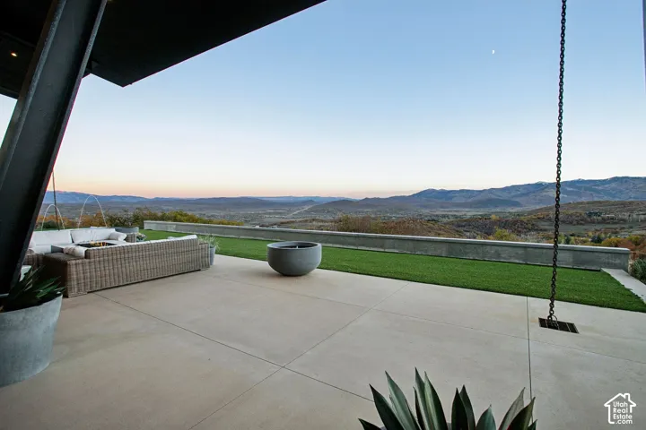 Covered concrete patio featuring a linear low-profile retaining wall, level synthetic turf lawn, metal chain downspout, and distant mountain backdrop
