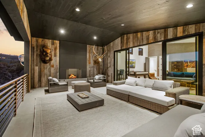 Covered outdoor living space featuring reclaimed wood wall paneling, matte black ceiling finish, recessed lighting, and a built-in fireplace with large-format tile surround, finished with a horizontal slat railing