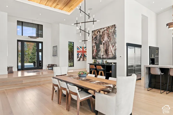 High-ceiling interior featuring a recessed wood-paneled tray ceiling, linear multi-arm chandelier, wood-finish flooring, bar, and black-framed glass entry door