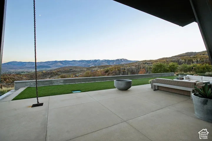 Covered concrete patio featuring a linear stone retaining wall, synthetic lawn, metal chain rain gutter, and mountain backdrop