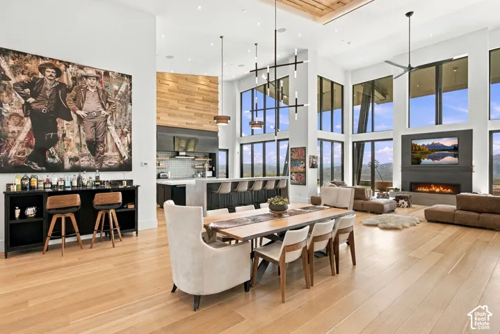 Open-concept great room featuring high ceilings with wood-plank detailing, floor-to-ceiling windows, wood-finish flooring, a linear fireplace with custom surround, and bar connected to designer kitchen