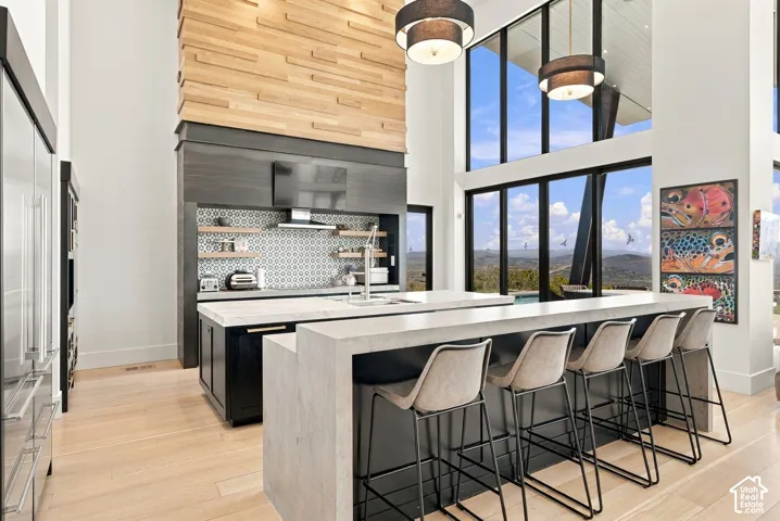 Double-height ceilings with floor-to-ceiling windows, central kitchen island with waterfall countertop, geometric patterned backsplash, wood-finish flooring, and tiered wood accent wall feature