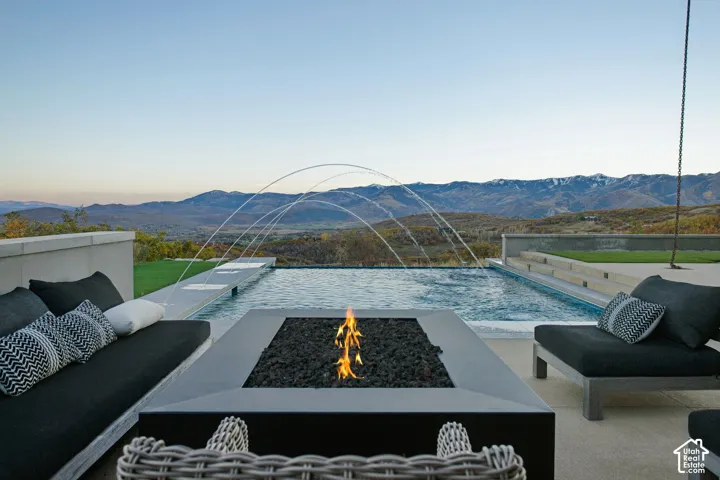 Concrete terrace featuring a built-in fire feature, an infinity-edge pool with water arches, and a mountain backdrop