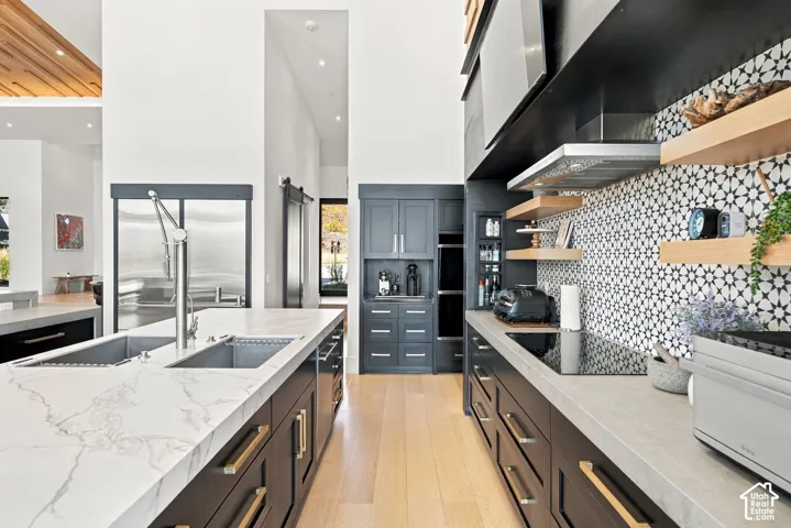 Kitchen featuring wood-finish flooring, stone-topped island with an integrated sink, geometric tile backsplash, built-in wall oven, and matte black cabinetry with brushed metal hardware