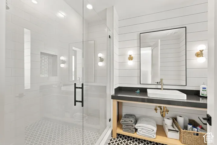 Glass-enclosed shower with tiled walls and built-in niche, white shiplap wall paneling, open-shelf wood vanity with dark countertop, rectangular vessel sink, and dual-globe brass wall sconces