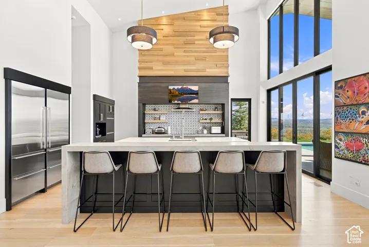 Vaulted ceilings featuring a floor-to-ceiling wood-paneled accent wall, expansive multi-pane windows, central island with a waterfall-edge countertop, stainless steel refrigerator, and recessed lighting