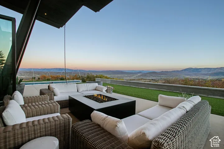 Covered concrete patio featuring a square built-in fire pit, synthetic lawn area, and stone retaining wall, with a backdrop of distant mountain ranges and a rectangular swimming pool