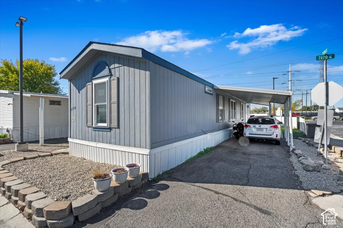 Single-story exterior featuring vertical light grey siding, a front-facing arched window, decorative shutters, and an attached covered carport