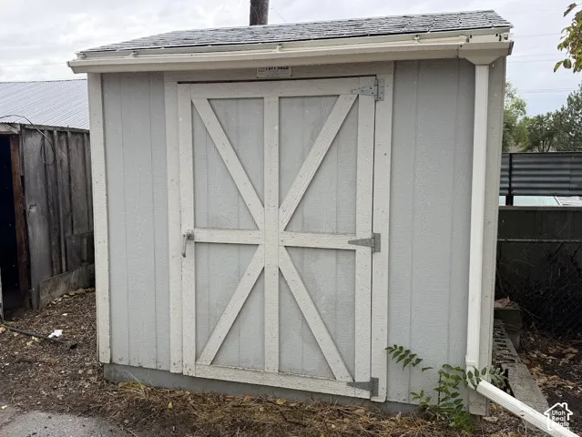 Detached storage structure with gray wood-paneled siding, cross-buck double doors, asphalt shingle roof, and integrated rain gutter system