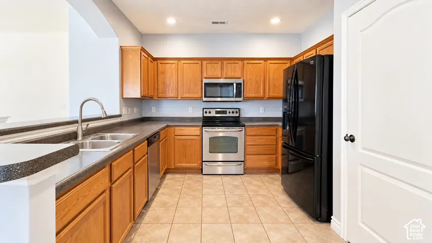 Kitchen featuring wooden cabinetry, dark countertops, tile flooring, stainless steel appliances, and a double-basin sink with a gooseneck faucet