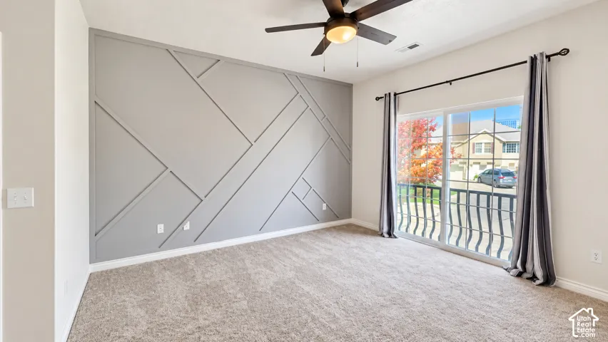 Geometric accent wall, carpeted flooring, ceiling fan with integrated lighting, and sliding glass door with a metal balcony railing