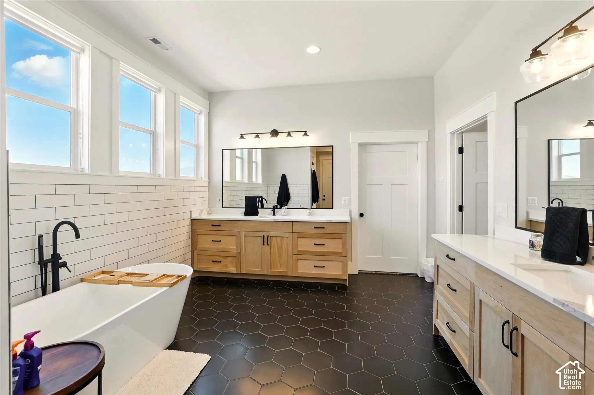 Primary bathroom with freestanding soaking tub with matte black faucet, dual wood-finish cabinetry with white stone countertops, subway tile wall treatment, and black hexagonal floor tiling