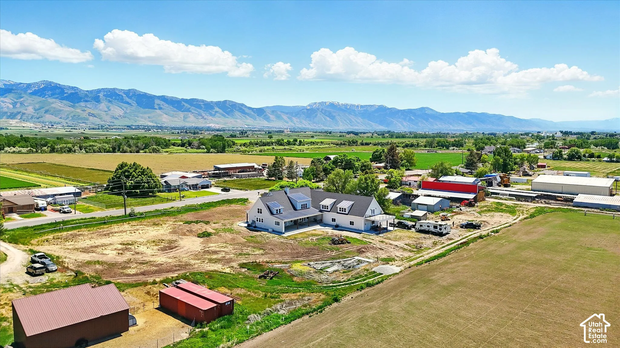 Aerial perspective of a multi-structure property featuring a large residence with a metal roof and dormer windows, two detached outbuildings, expansive acreage, and a prominent mountain backdrop