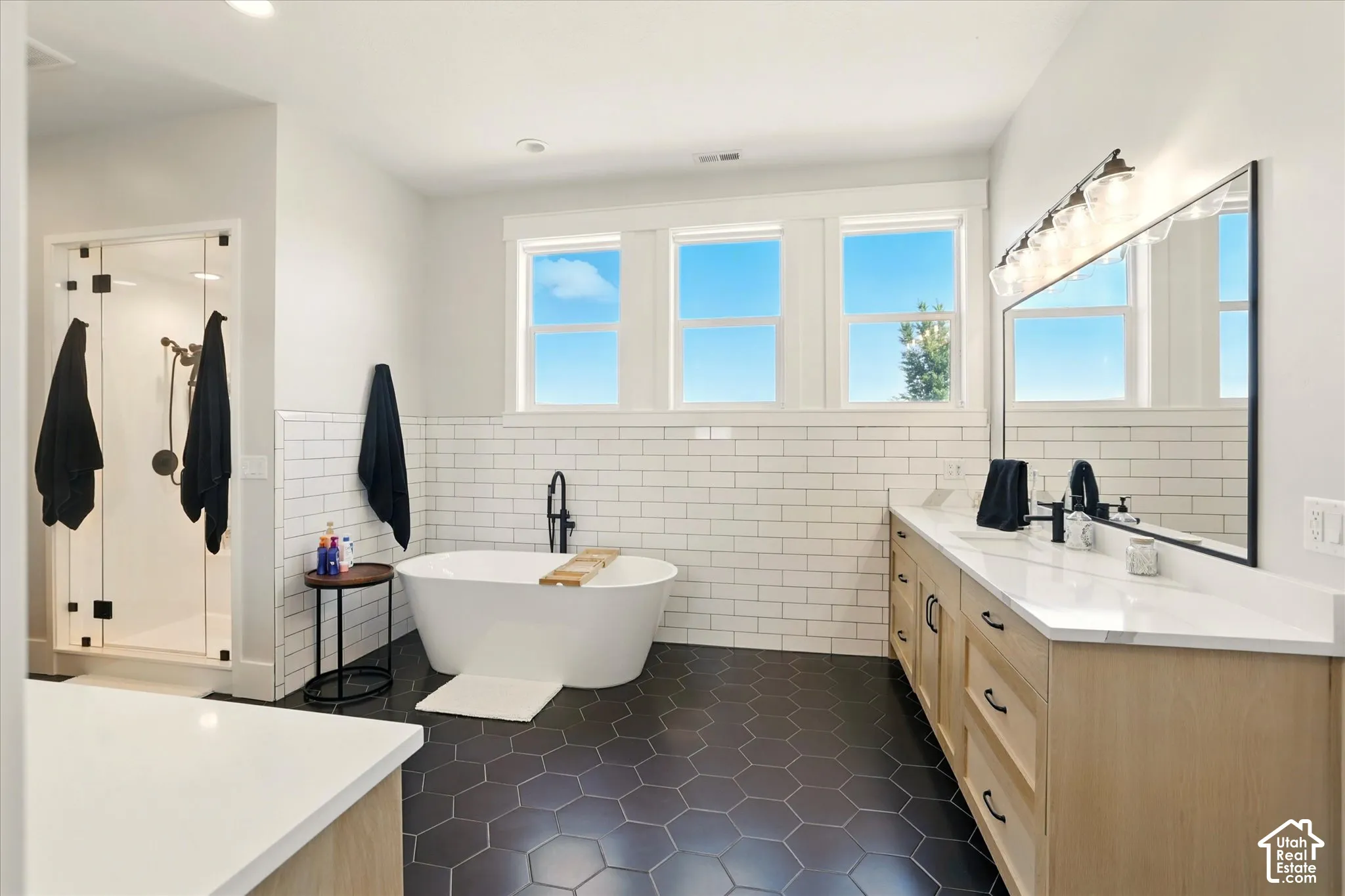 Primary bathroom with freestanding soaking tub with a floor-mounted matte black faucet,  vanity with stone countertops, glass-enclosed shower, subway tile wall accents, and hexagonal floor tiling