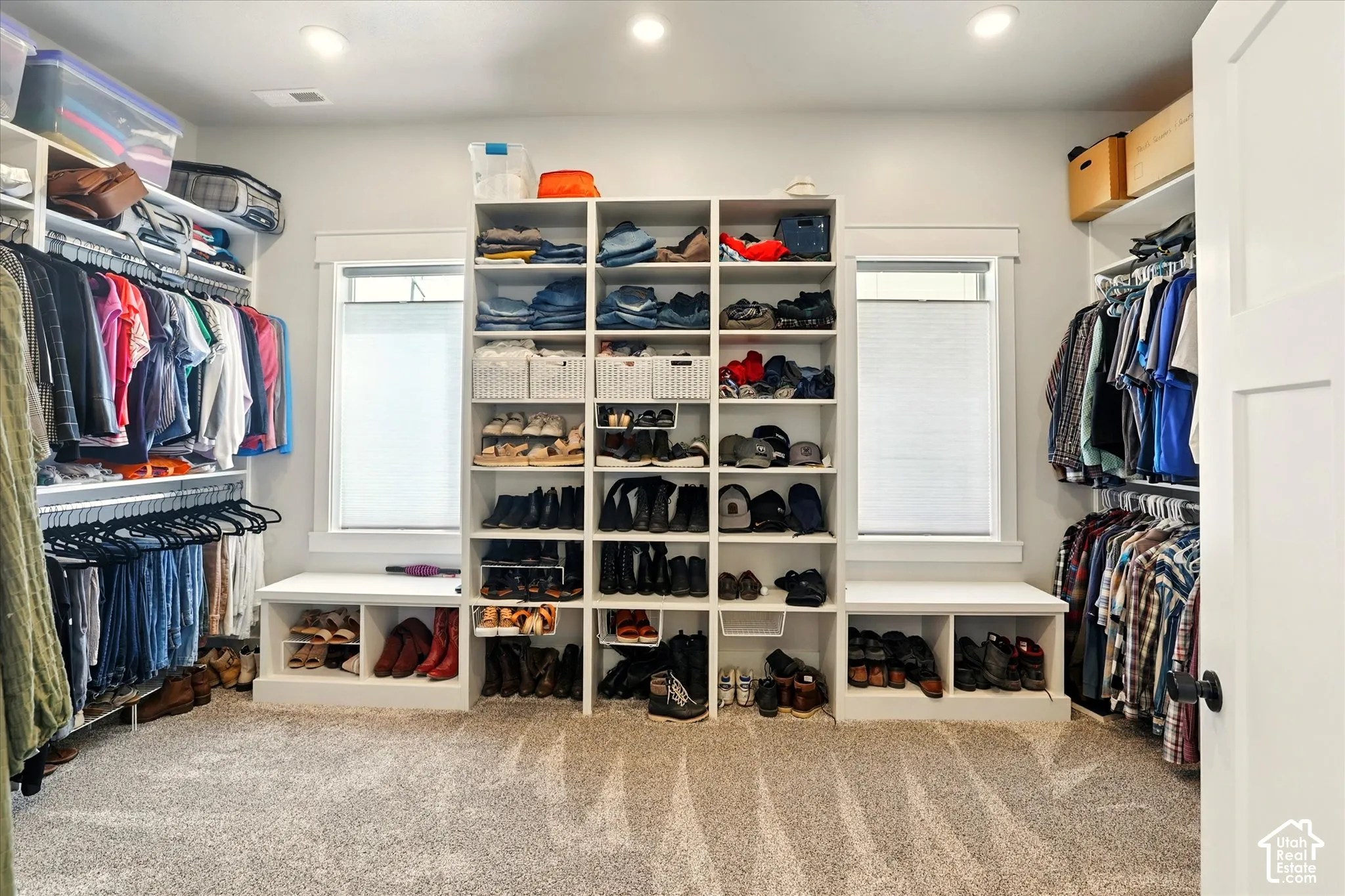 Custom walk-in closet featuring floor-to-ceiling built-in shelving, integrated lower storage benches, dual windows with casing, recessed lighting, and carpeted flooring