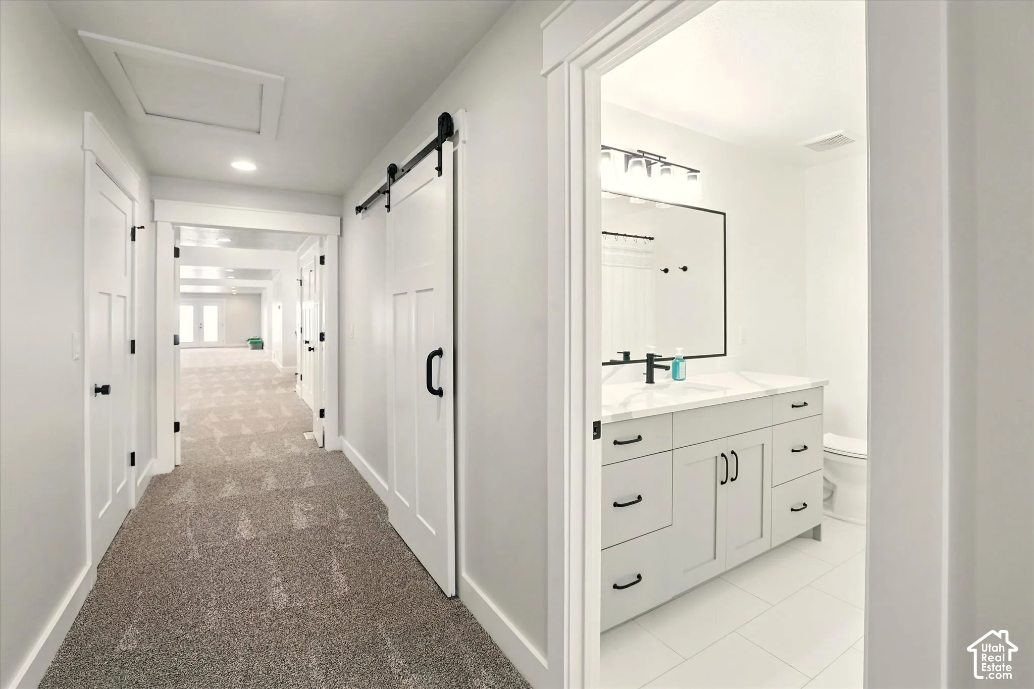 Hallway featuring carpeted flooring and a sliding barn door to closet, a bathroom with a white vanity, stone-finish countertop, black hardware, and large rectangular mirror