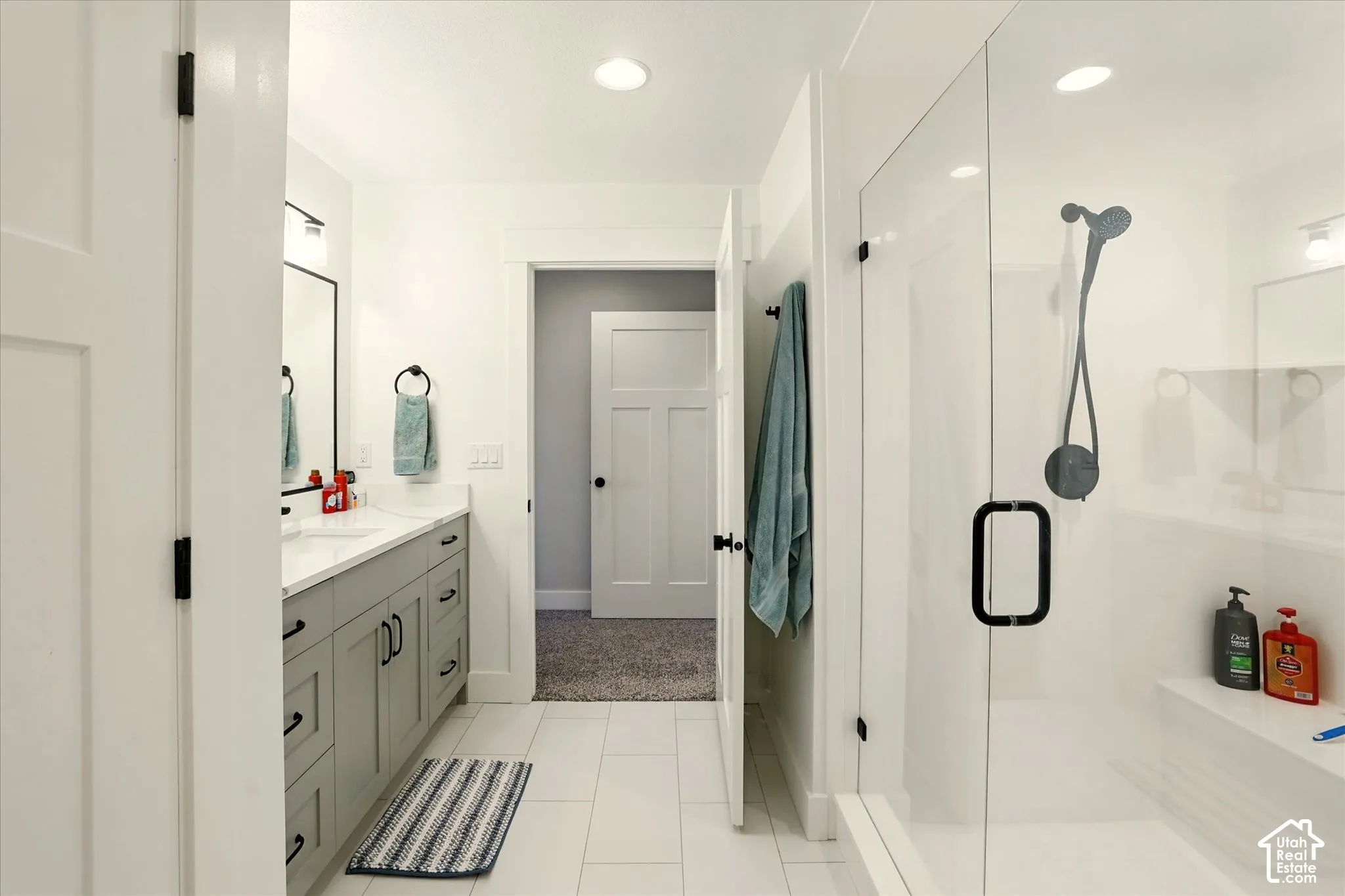 Bathroom featuring a walk-in shower with glass enclosure, light gray shaker-style cabinetry, solid-surface countertop, matte black hardware, and large-format tile flooring