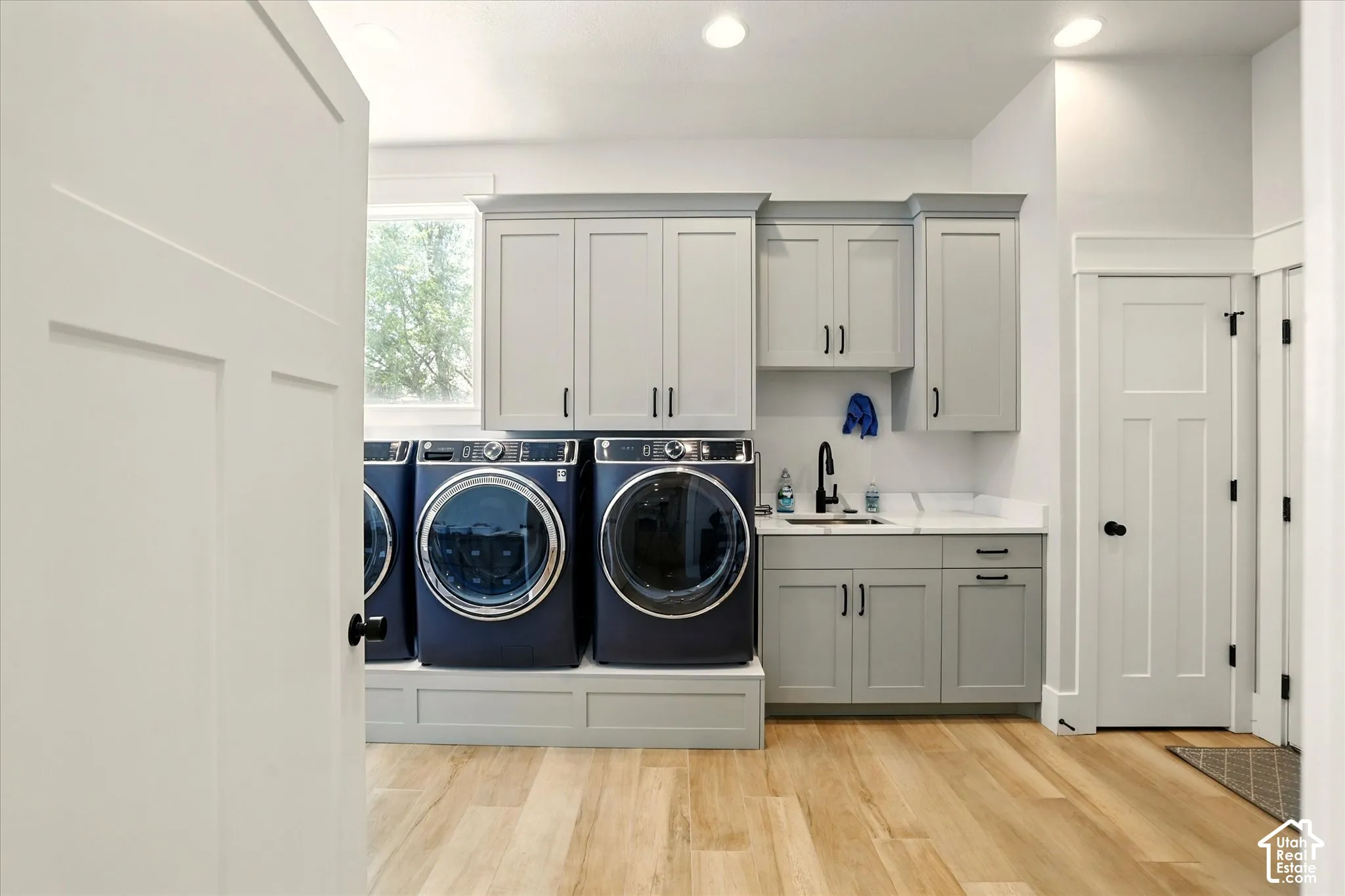 Laundry room featuring light grey shaker-style cabinetry, white stone-finish countertops, an undermount sink with a matte black faucet, elevated appliance platform, and wood-finish flooring