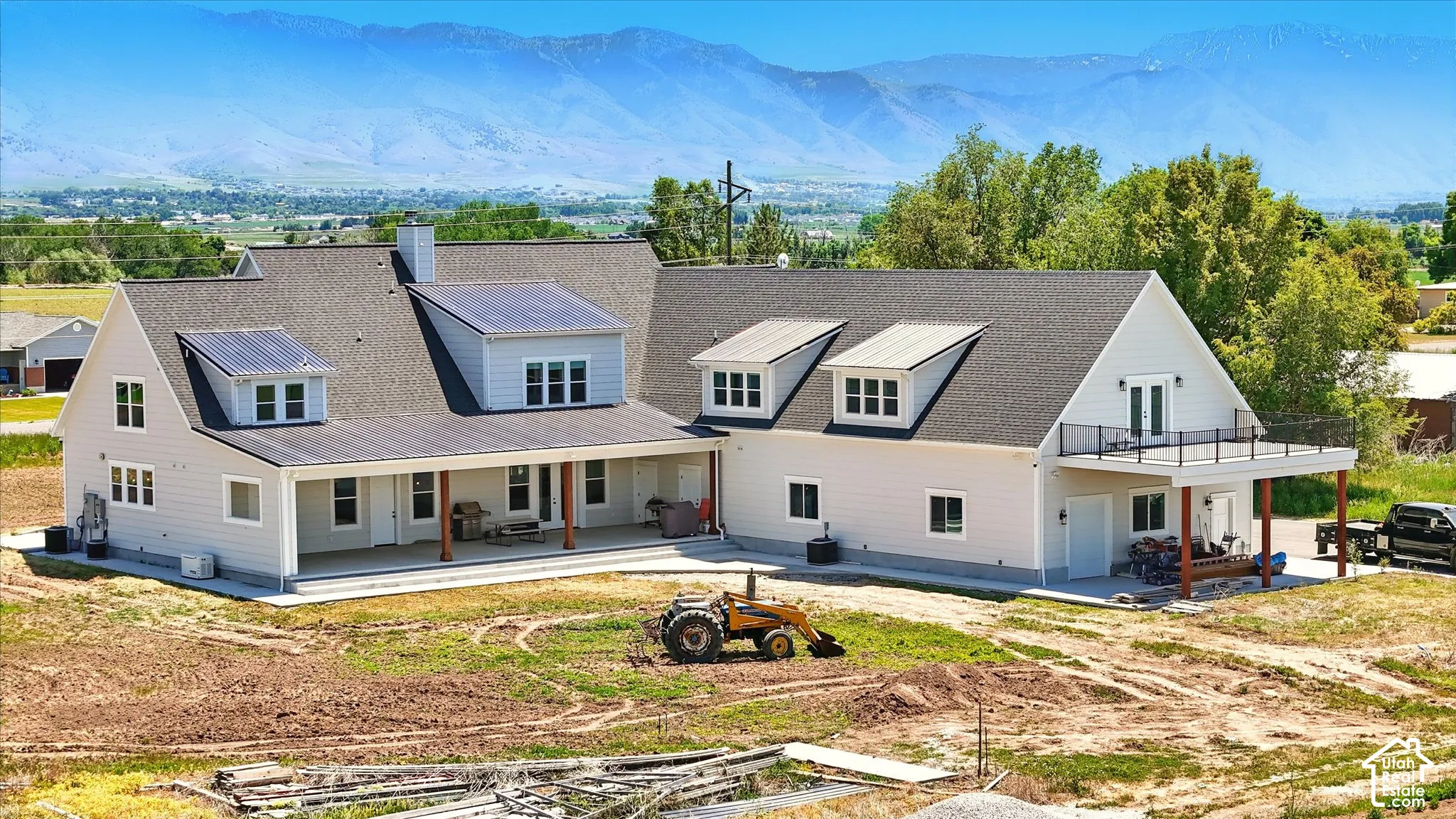 Exterior featuring light-toned horizontal siding, expansive covered patio with wooden support columns, second-story balcony with metal railing, and mountain backdrop