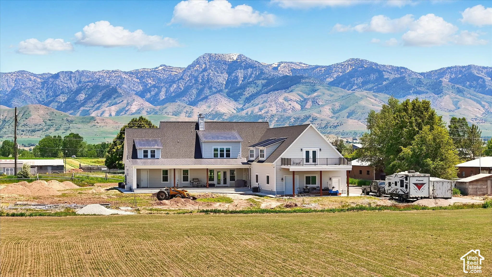 Multi-level exterior featuring light-toned siding, a shingled roof with metal roof on dormers and covered patio, a second-story balcony with metal railings, and prominent mountain backdrops