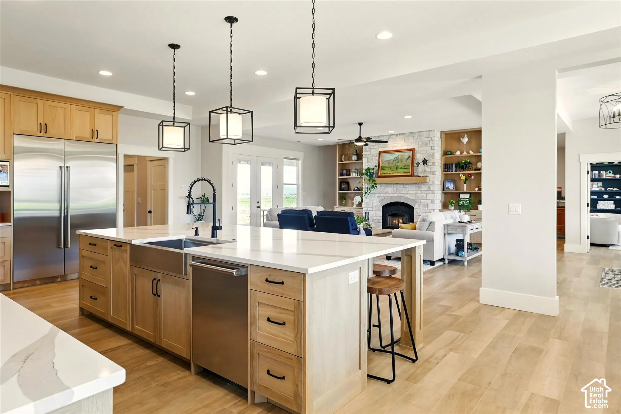 Kitchen featuring a central island with a quartz countertop, stainless steel apron-front sink, wood-finish cabinetry, and industrial-style pendant lighting, complemented by a floor-to-ceiling stone fireplace with built-in shelving