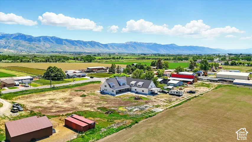 Aerial perspective of a multi-structure property featuring a large residence with a metal roof and dormer windows, two detached outbuildings, expansive acreage, and a prominent mountain backdrop