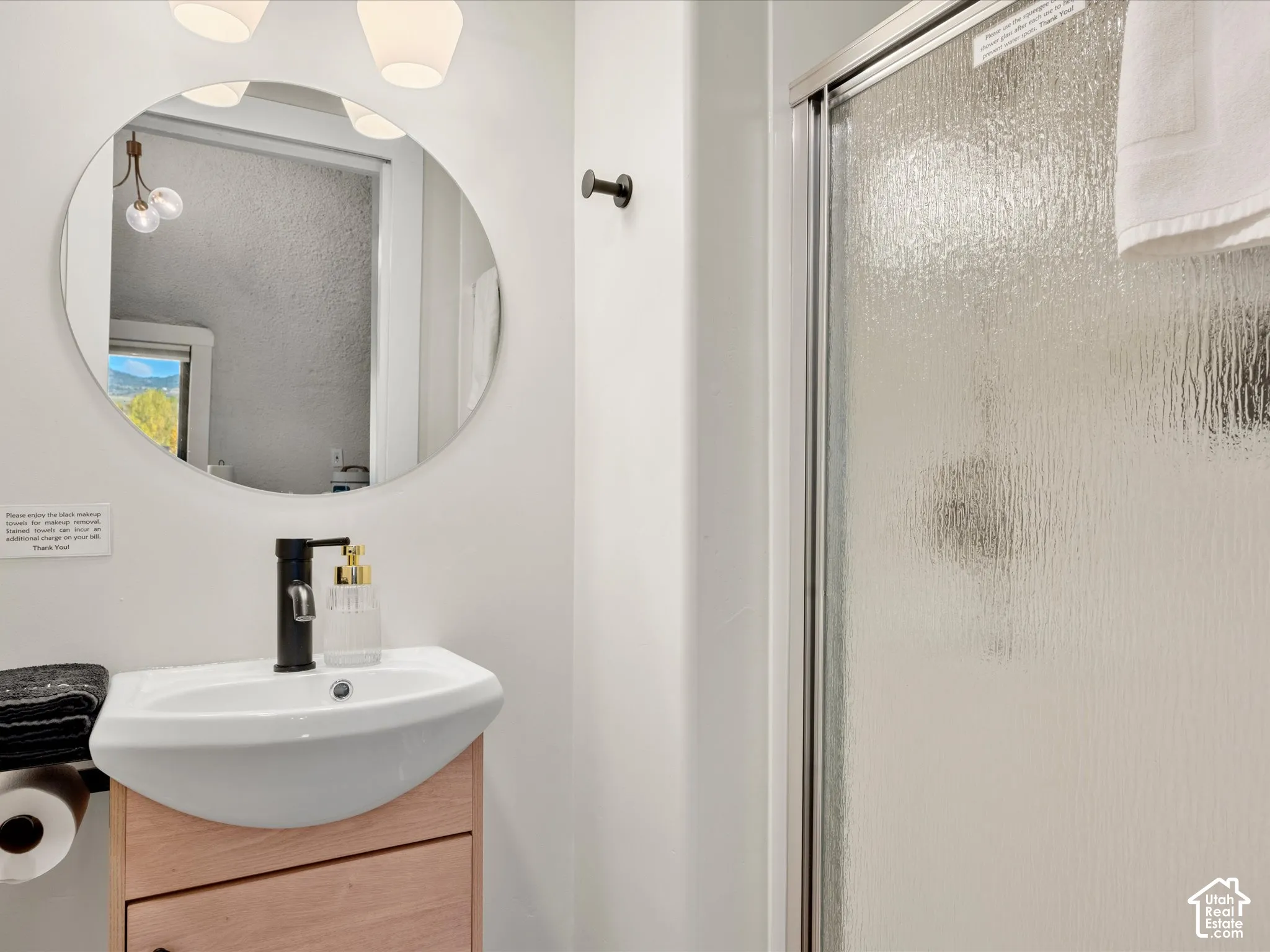 Wall-mounted circular mirror, vanity with wood-finish cabinetry, matte black faucet, and walk-in shower with textured glass enclosure