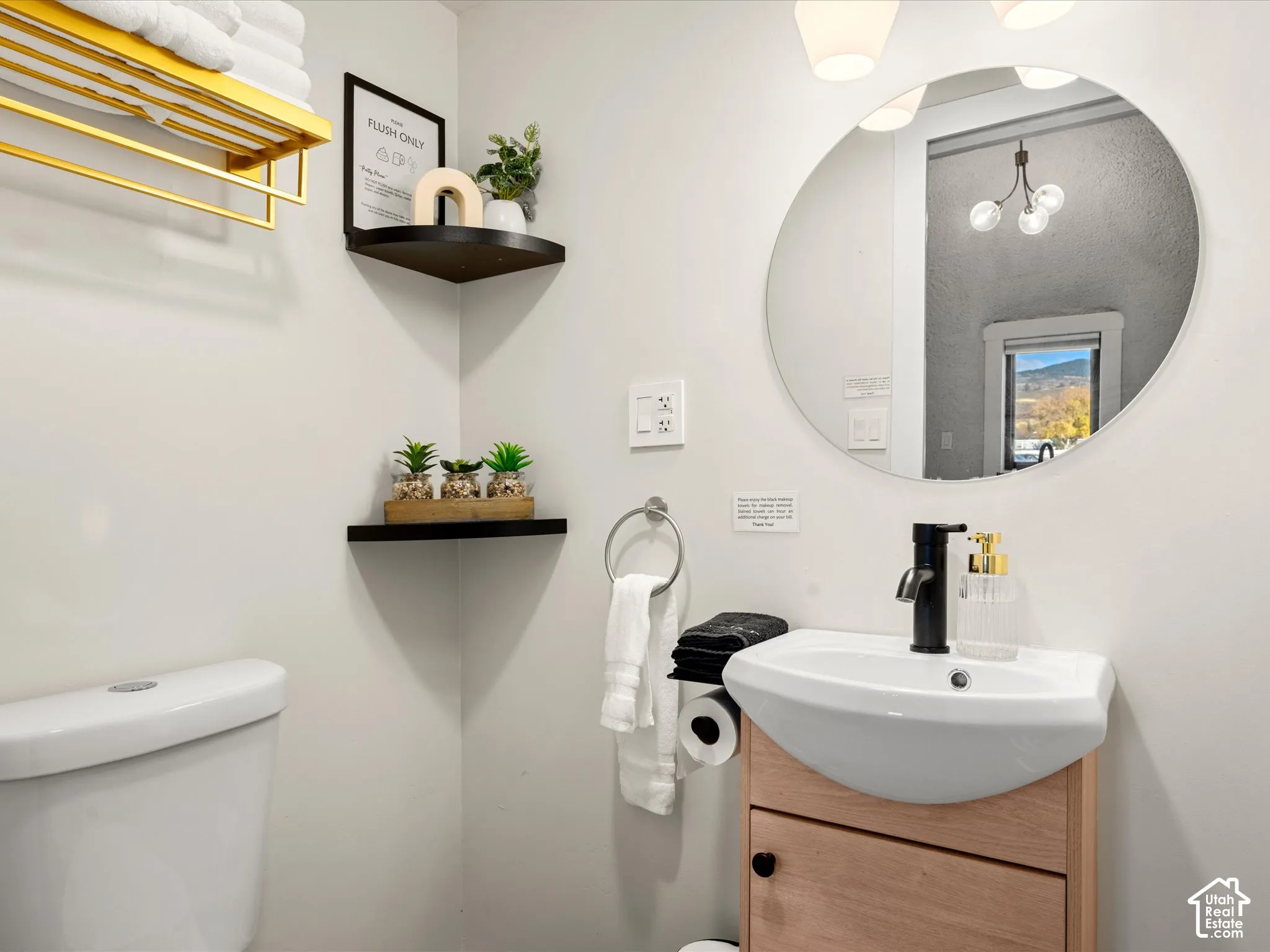 Wall-mounted gold-tone towel rack, circular frameless mirror, wood-finish vanity with a ceramic basin, matte black faucet, and built-in corner shelving