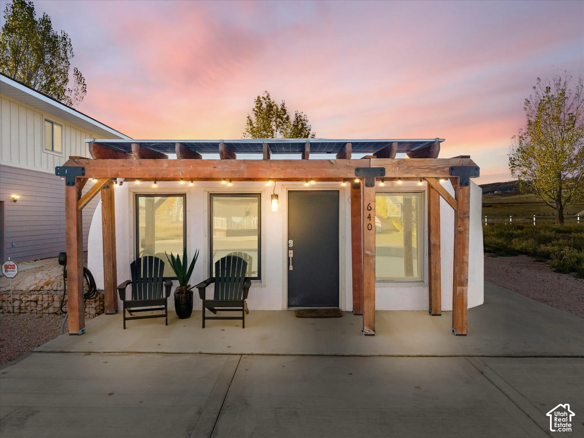 Exterior facade featuring a timber-frame pergola, integrated roof-mounted solar panels, concrete porch, and three large rectangular windows