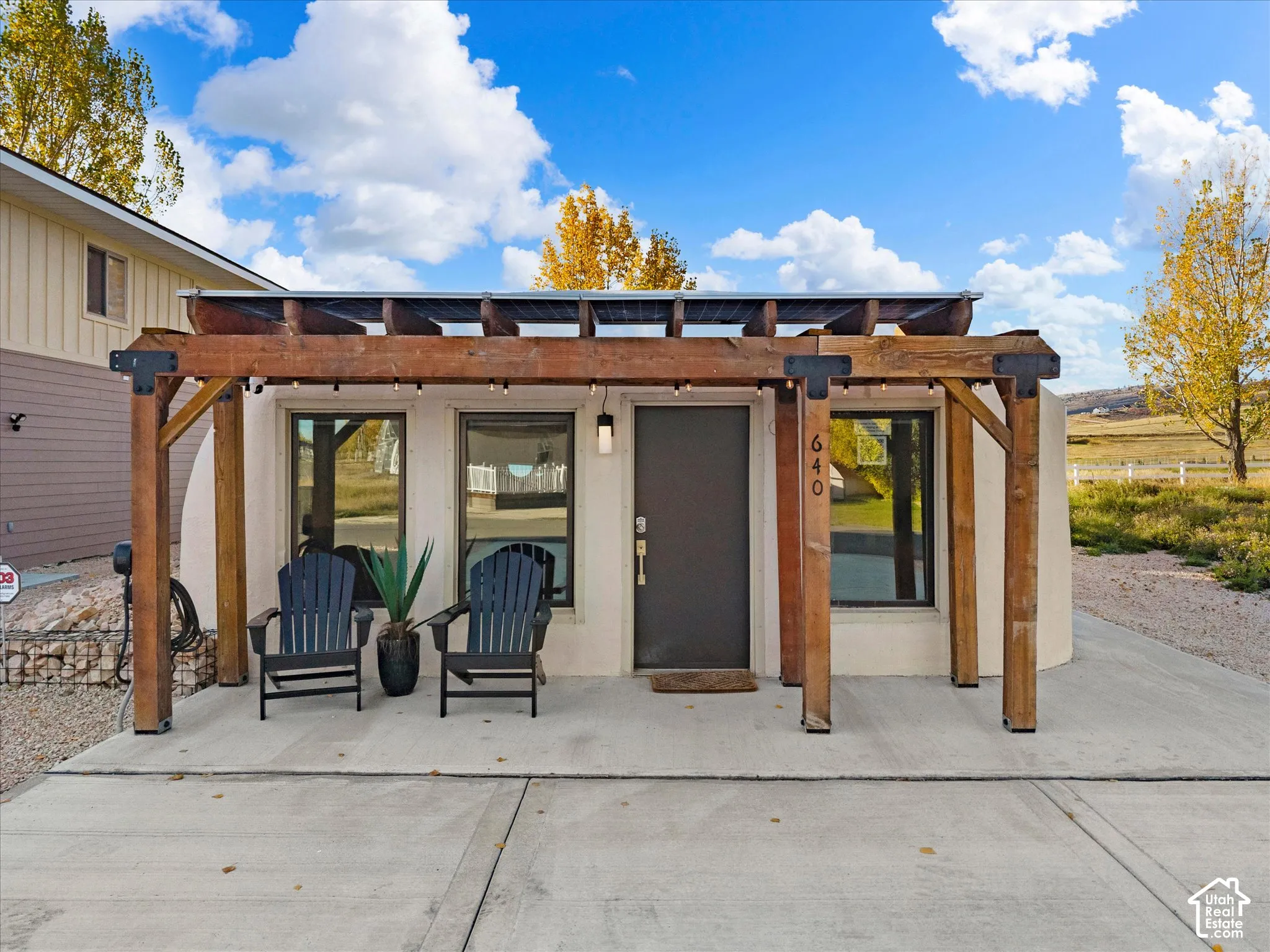 Exterior entrance featuring a concrete patio, wood-beam pergola with metal hardware, smooth-finish siding, and rectangular windows