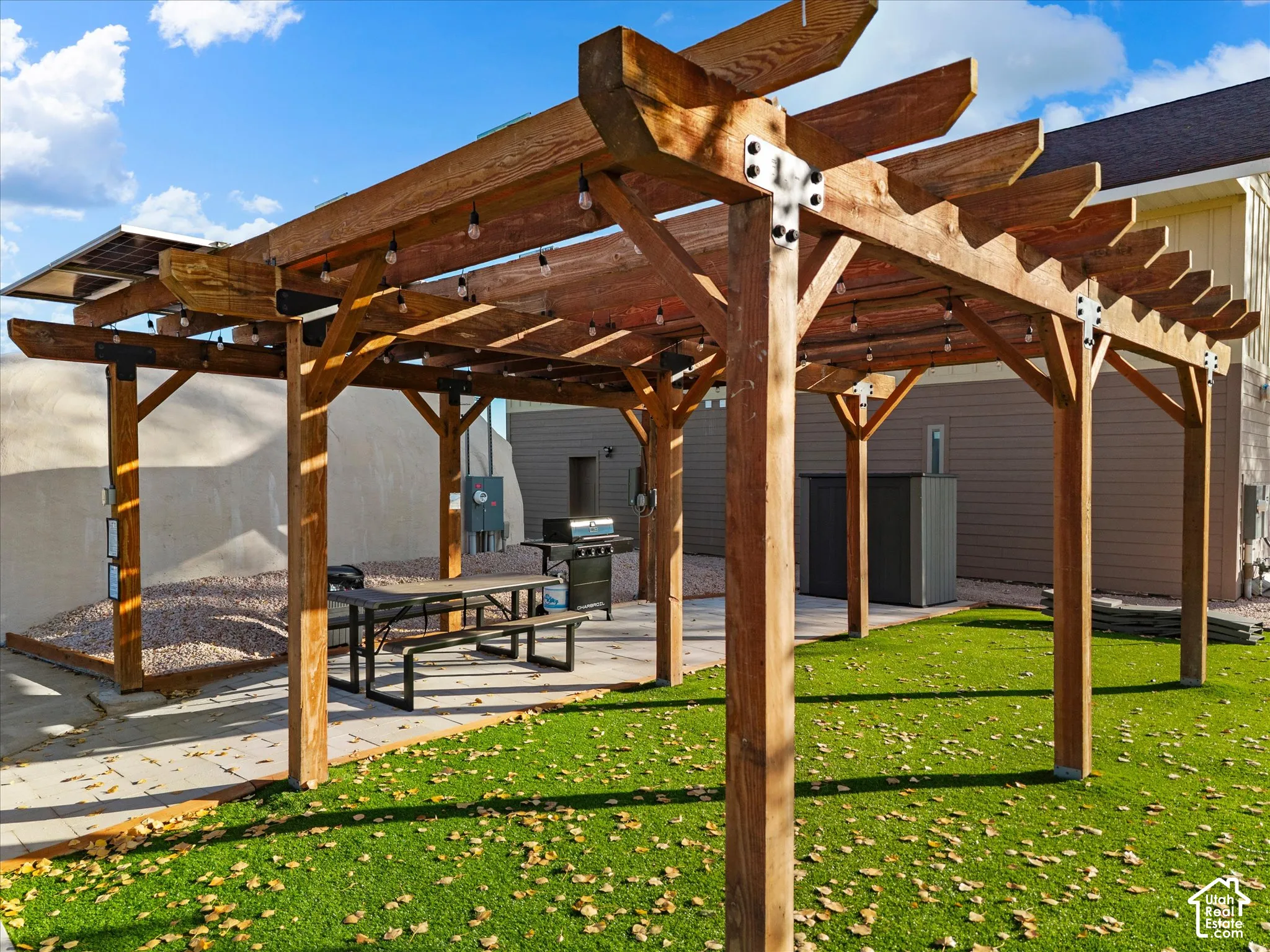 Custom wood-frame pergola with metal mounting hardware, stone-paver patio, artificial turf lawn, and integrated solar panels
