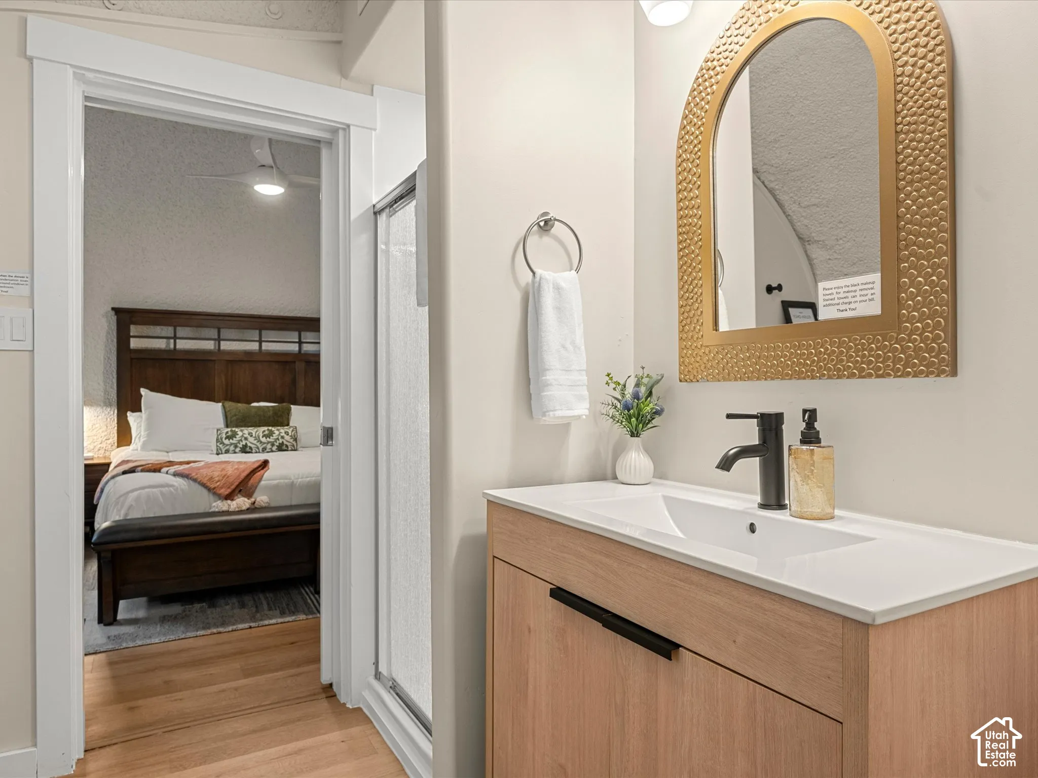 Bathroom vanity featuring a light wood-finish cabinet, integrated white sink, and matte black faucet hardware, complemented by an arched decorative mirror and wood-finish flooring