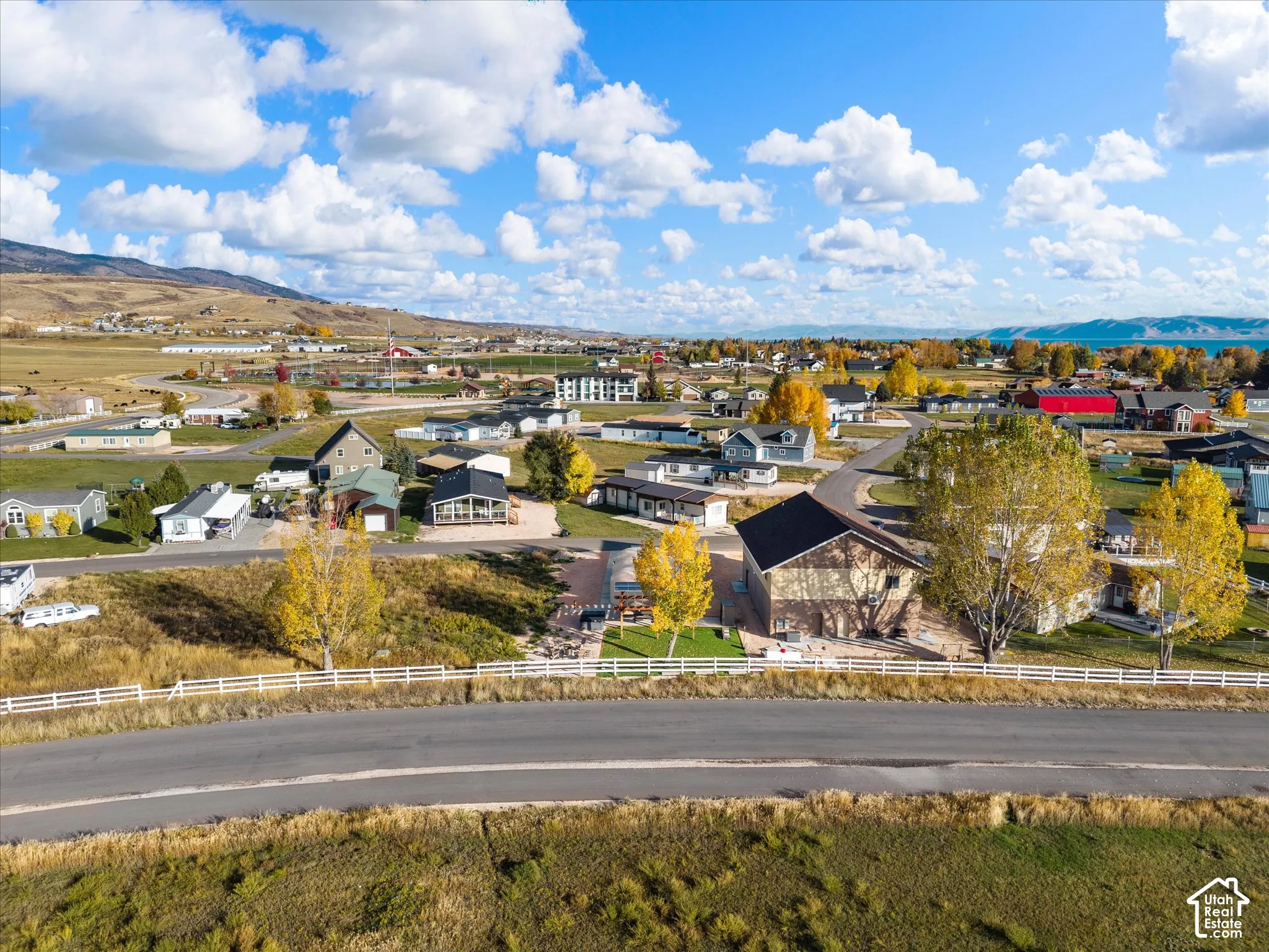 Aerial perspective featuring a multi-story residence with a paved driveway, white perimeter fencing, and established trees set against a prominent mountain and water landscape