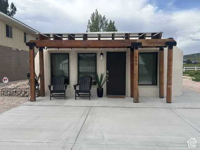 Custom wood pergola entrance featuring exposed structural beams with dark metal accents, smooth stucco facade, concrete slab patio, and large glass windows