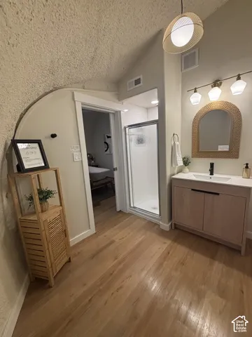 Bathroom featuring wood-finish flooring, a wood-toned vanity with a white countertop, a walk-in shower with a glass enclosure, an arched wall mirror, and dual-style lighting fixtures