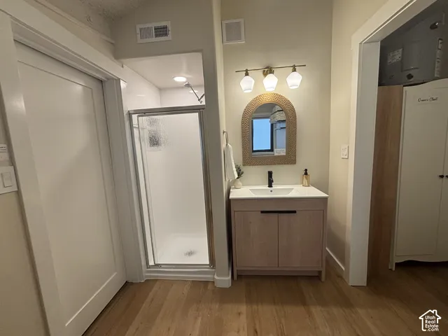 Bathroom featuring a wood-finish vanity with a white countertop and integrated sink, a glass-enclosed shower, a wall-mounted three-light fixture, and wood-finish flooring