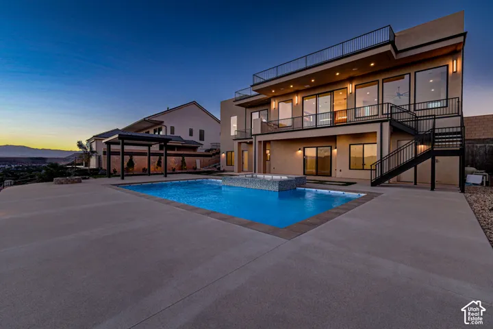 In-ground swimming pool with an integrated spa, expansive concrete patio, detached pergola, multi-level balconies with metal railings, and exterior staircase