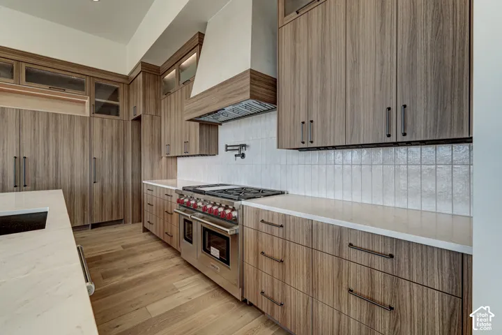Kitchen featuring custom wood-finish cabinetry, a stainless steel Wolf range, a white tile backsplash, light-toned countertops, and wood-finish flooring