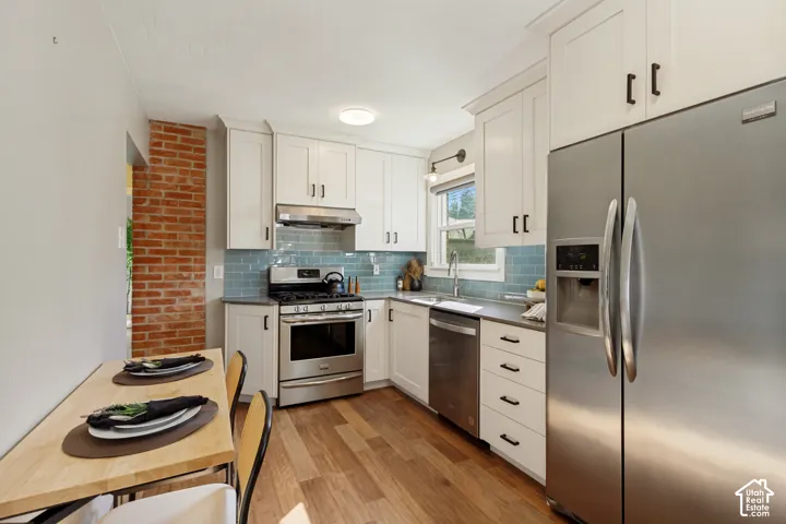 Kitchen featuring shaker-style cabinetry, stainless steel appliances, subway tile backsplash, exposed brick accent wall, and wood-finish flooring