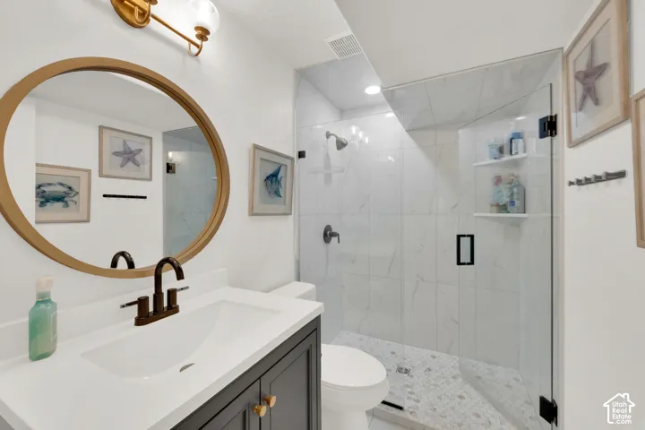 Bathroom featuring a vanity with white countertop, circular gold-toned mirror, glass-enclosed shower with tiled surround, stone-tiled floor, and dark bronze-toned fixtures