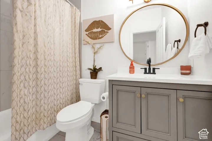 Grey-toned shaker vanity with a white countertop, round gold-rimmed mirror, matte black faucet, and a shower-tub combination with light-toned tile surround